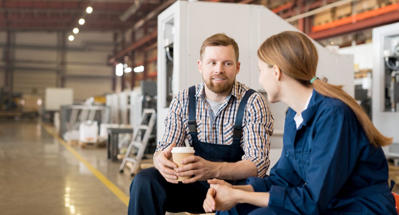 Two colleagues discussing in a grey board production workshop one holding a hot drink highlighting collaboration in eco friendly packaging manufacturing