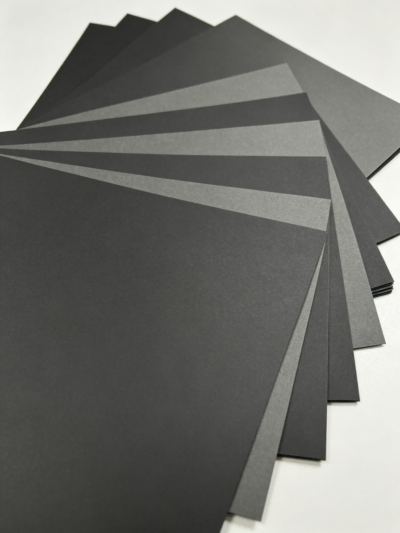 Single‑Sided Through‑Black Paperboard fan layout, assorted black & dark grey matte cardboard sheets flat lay, solid core black paper board sample set