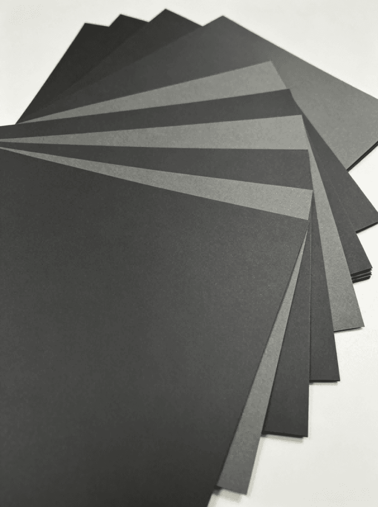 Single‑Sided Through‑Black Paperboard fan layout, assorted black & dark grey matte cardboard sheets flat lay, solid core black paper board sample set